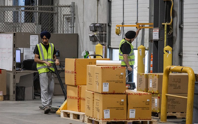 Warehouse workers moving boxed orders on pallet jacks near a loading dock, representing QRC Logistics ecommerce fulfillment services. Warehouse workers moving boxed orders on pallet jacks near a loading dock, representing QRC Logistics ecommerce fulfillment services.