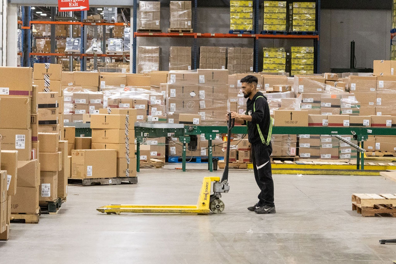 Warehouse associate moving an empty pallet jack past staged pallets and a conveyor line at QRC’s warehouse in Halton Hills